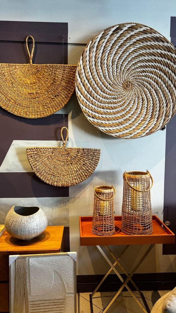 Peças de cestaria penduradas na parede, incluindo cestas de fibras naturais em formatos circulares e semicirculares, complementadas por lanternas de vime sobre uma mesa decorativa ao lado de vasos de cerâmica e quadros minimalistas.