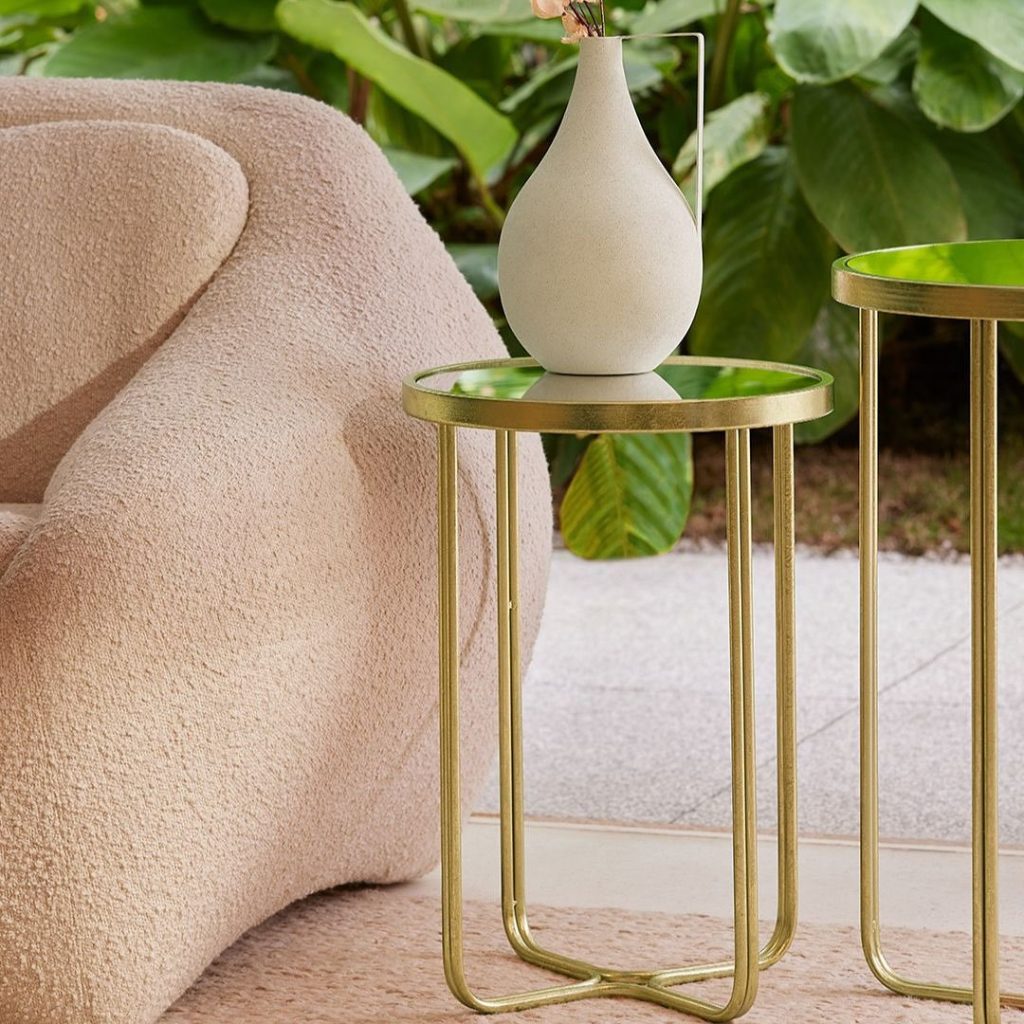 ar de mesas de canto douradas com tampo de vidro espelhado, decoradas com um vaso de cerâmica de design orgânico. A imagem ilustra a funcionalidade da mesa de canto como suporte para objetos decorativos.