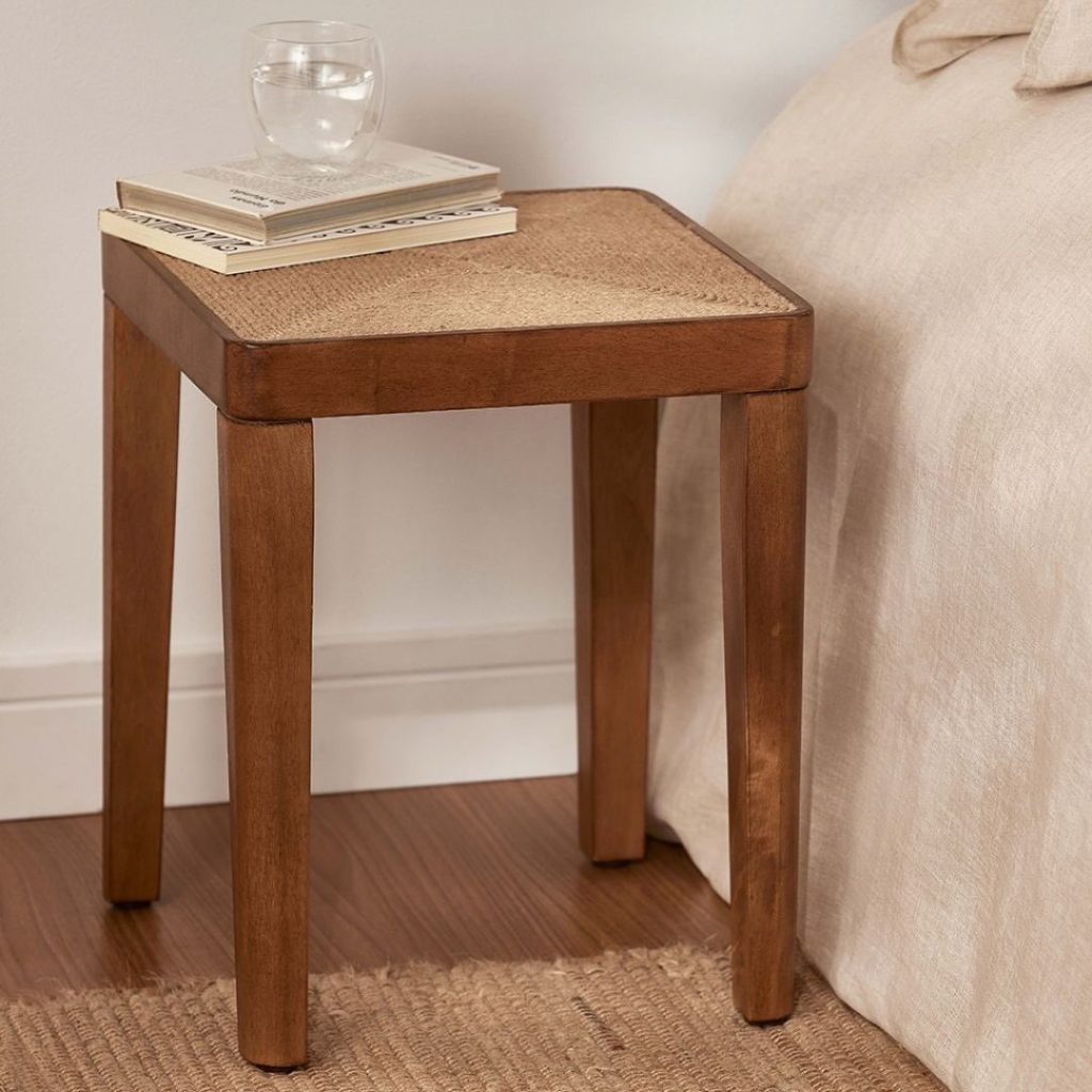 Mesa de canto de madeira com tampo de palha e design minimalista, decorada com livros e um copo de água, ao lado de uma cama. Perfeita para usar como mesa de cabeceira.