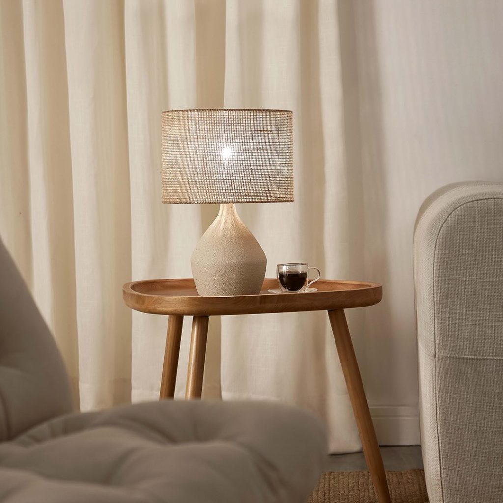 Abajur com cúpula de textura natural sobre uma mesa de canto de madeira com uma xícara de café, criando uma iluminação indireta e aconchegante para decoração de outono.