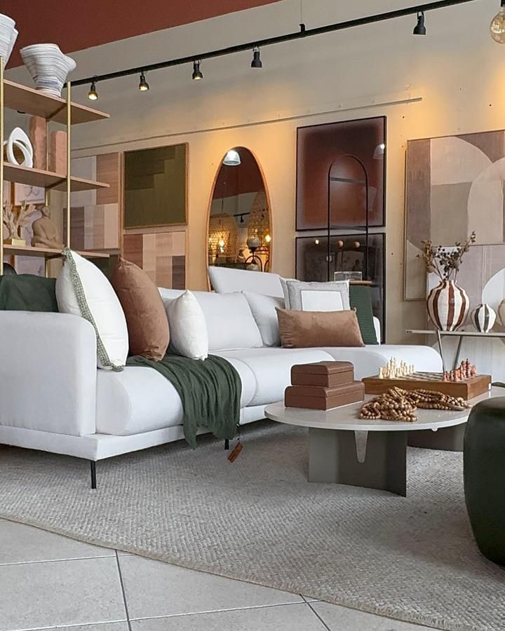 Ambiente de loja de decoração mostrando um sofá branco moderno decorado com almofadas terracota e verde oliva, manta verde e mesa de centro decorada com tabuleiro de xadrez em madeira.