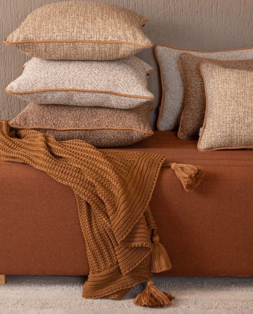 Pilha de almofadas sofisticadas em tons terrosos e uma manta de tricô terracota com franjas sobre o sofá, trazendo texturas quentes para os dias frios no Sul de Minas.